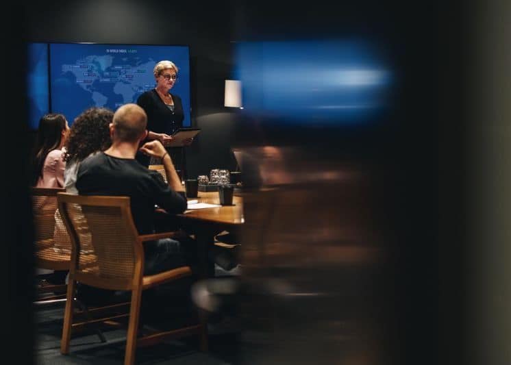 A group of people sitting around a large table in a concerence room. One person is holding a clipboard and presenting. In the background is a screen showing the global map.