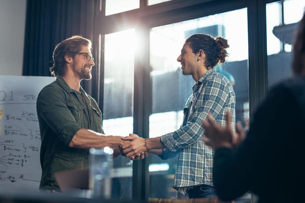 Two people shaking both hands in front of a whiteboard in a conference room, in a celebratory way. In the foreground, someone is clapping.