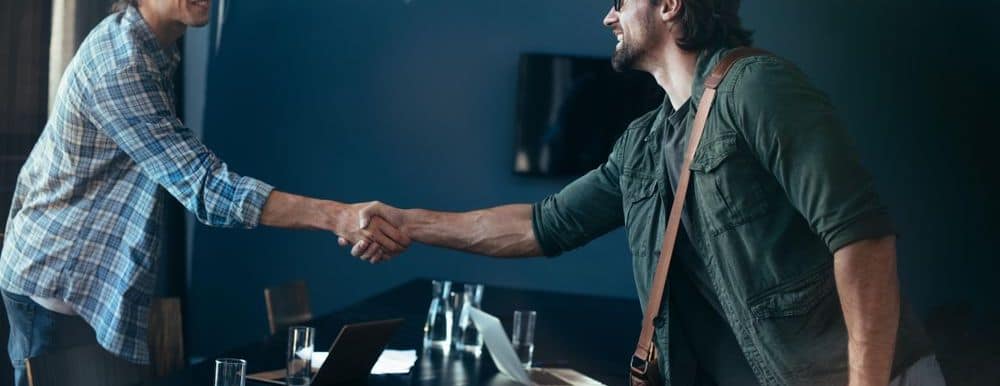 Two smiling, casually-dressed people are shaking hands over a large table in a meeting room. On the table are laptops and glasses of water.